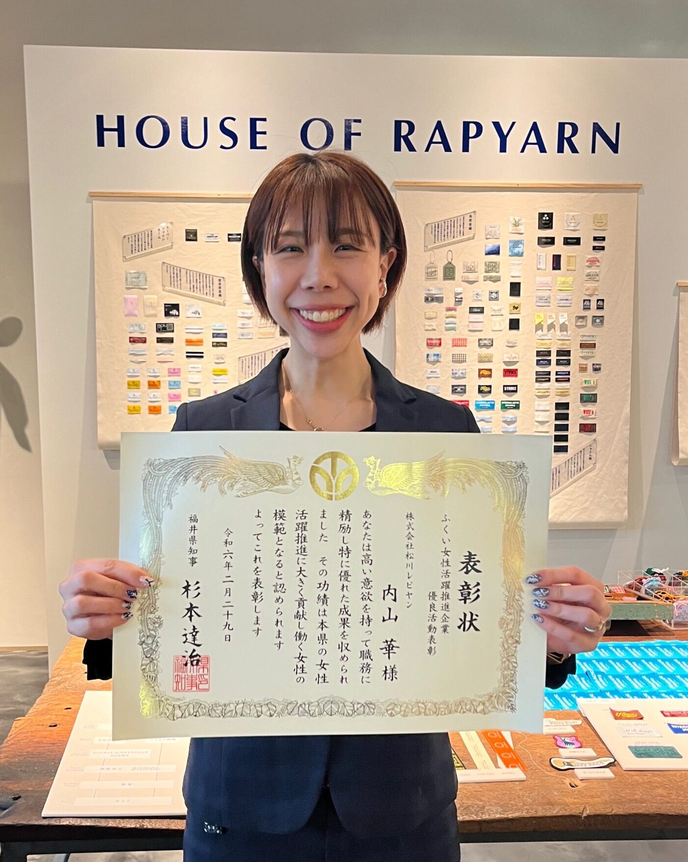 「HOUSE OF RAPYARN」の展示壁の前で、表彰状を掲げて笑う受賞した社員